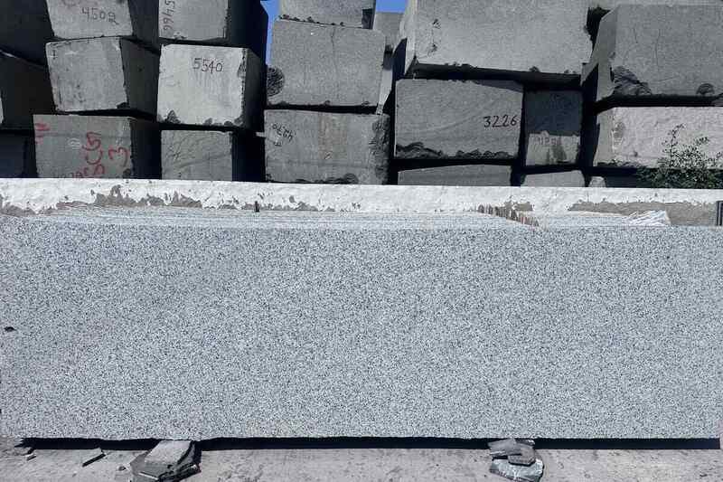 A large rectangular slab of Salt and Pepper granite featuring a light grey base with dense, fine black grains, speckles, and dot-like mineral inclusions; background shows stacked rough granite blocks.